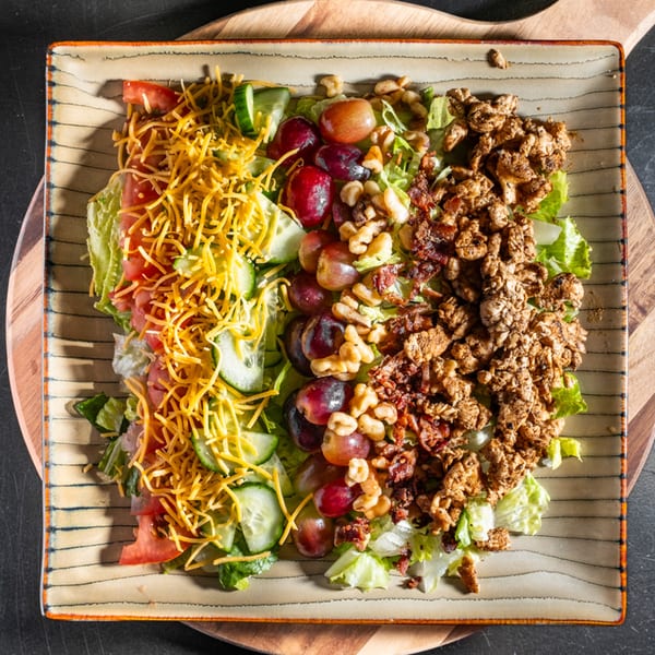 A colorful salad with shredded cheese, tomatoes, cucumbers, grapes, walnuts, and chicken on a square plate.