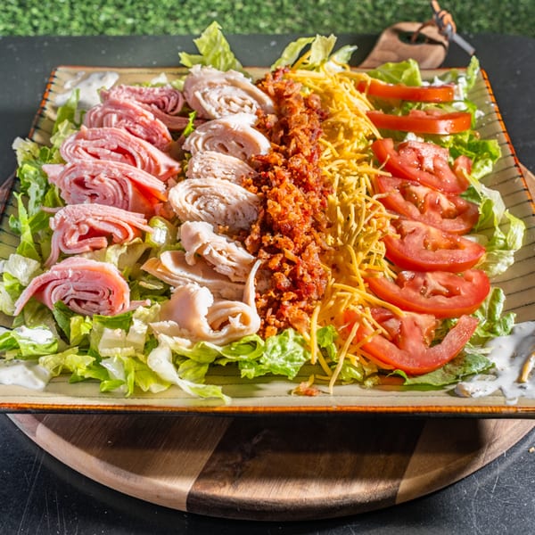 A large salad with ham, turkey, bacon bits, shredded cheese, tomatoes, and lettuce.